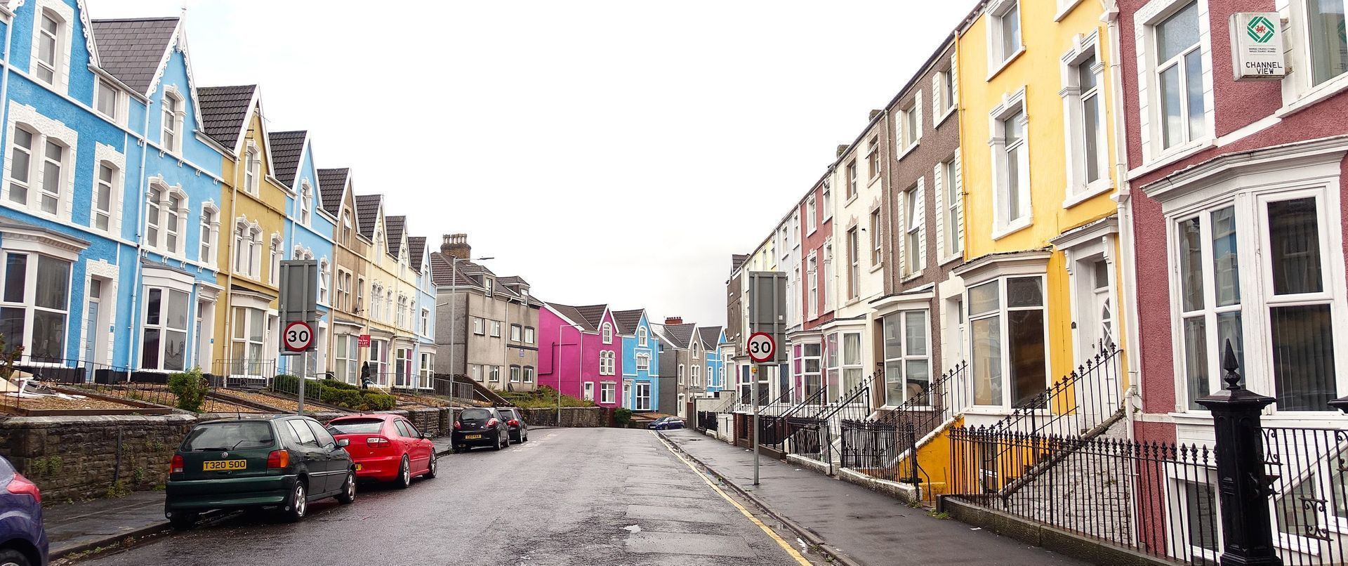 Colorful row houses line a street with parked cars, cloudy sky overhead.