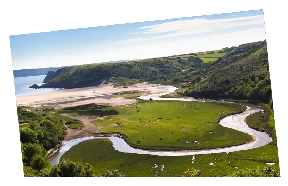A winding river flows to a beach, with green fields and hills in a scenic landscape.