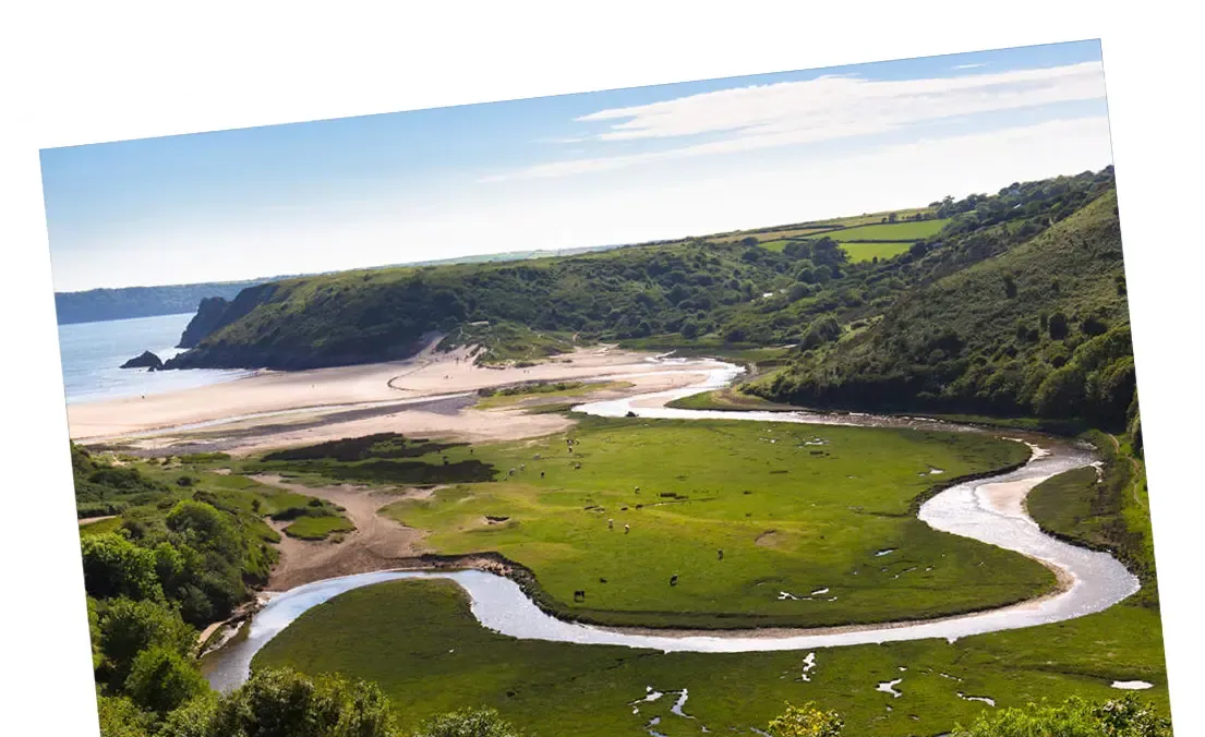 A winding river flows to a beach, with green fields and hills in a scenic landscape.
