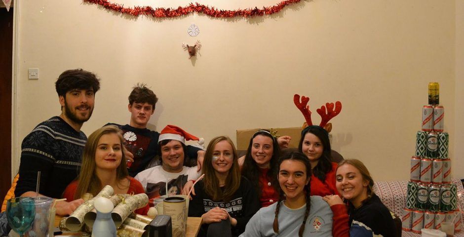 Group of friends at Christmas. Some wear festive sweaters and antlers, with a beer can Christmas tree.