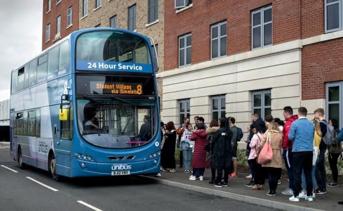 Blue double-decker bus at a bus stop, picking up passengers. 24-hour service displayed. Brick building in the background.