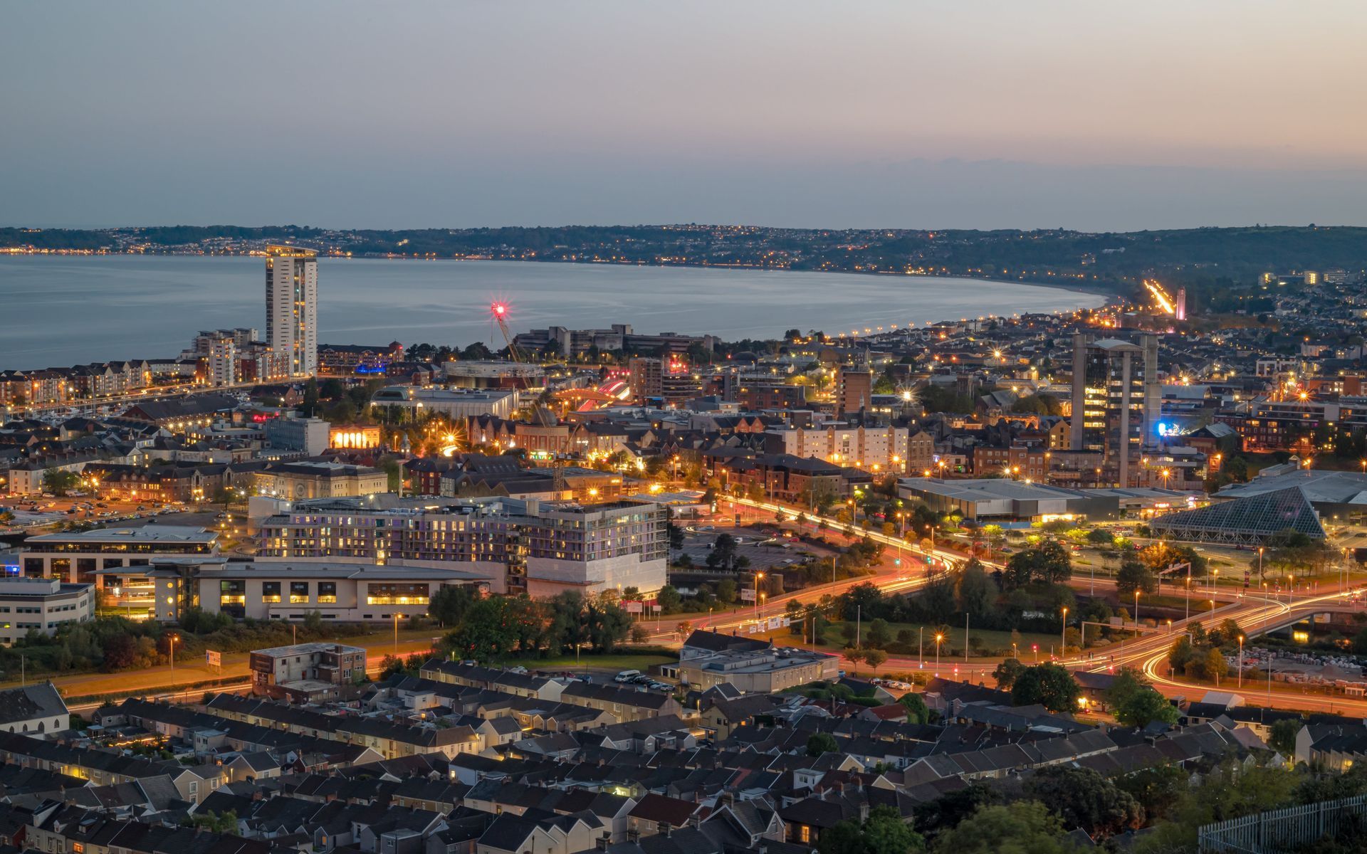 City at dusk, lights reflecting on water, tall buildings, roadways, and residential areas.