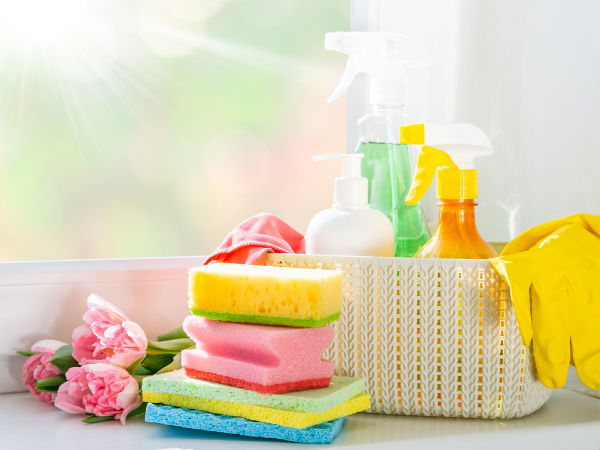 Cleaning supplies, including sponges, spray bottles, and gloves, sit on a windowsill next to tulips.