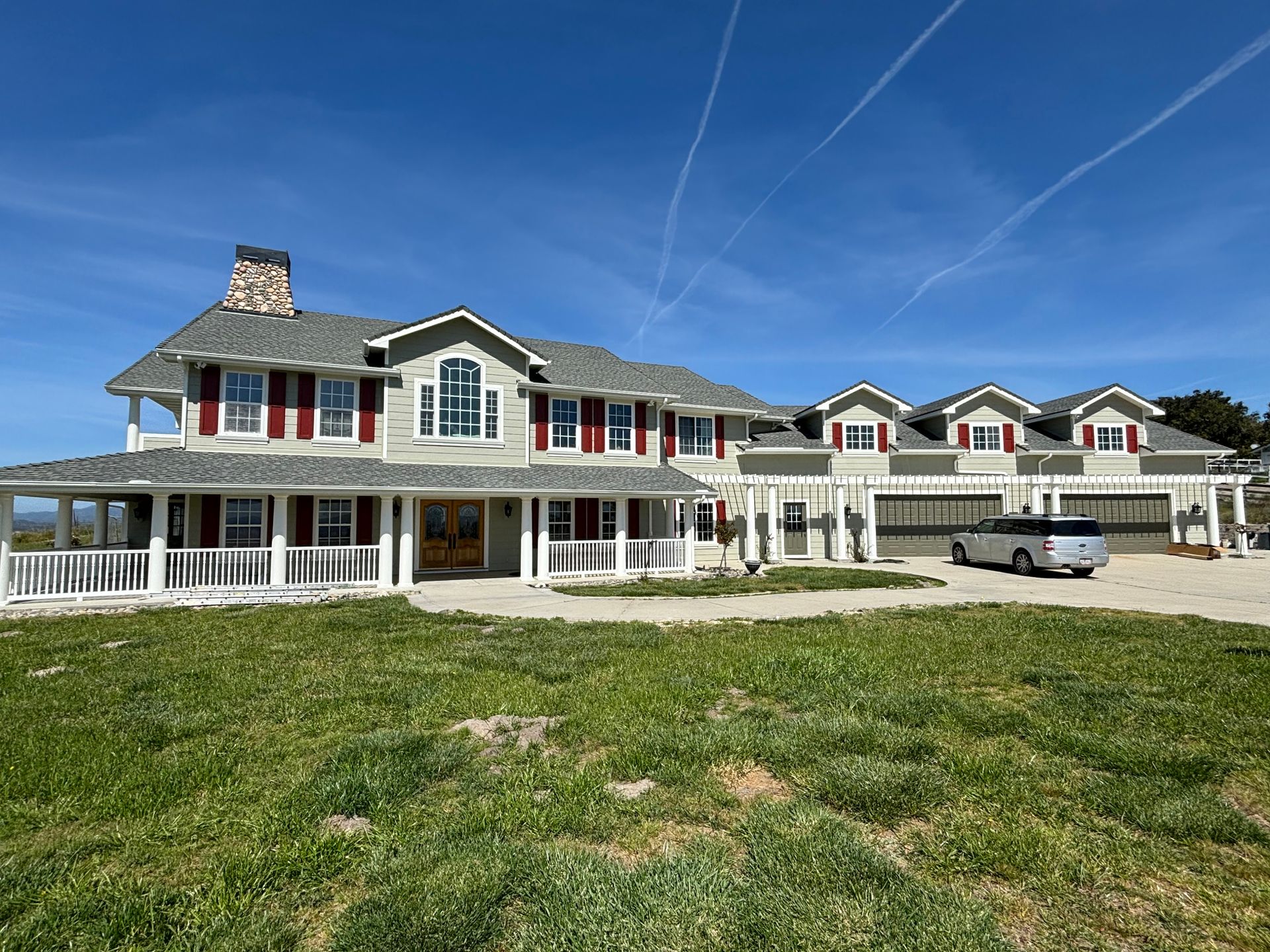 Large gray house with red shutters and a wrap-around porch, on a grassy plot under a blue sky.