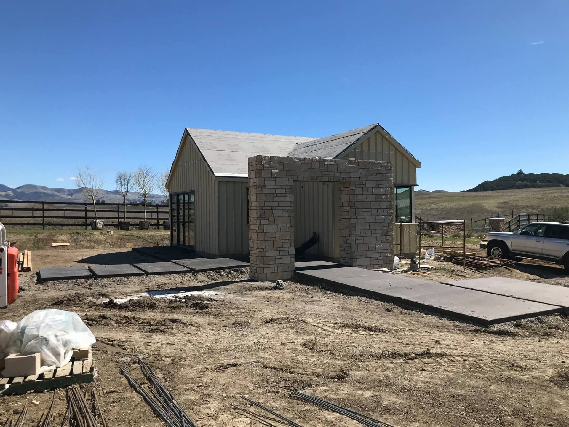 Building construction site with a light-colored wooden structure and a stone entryway under a blue sky.