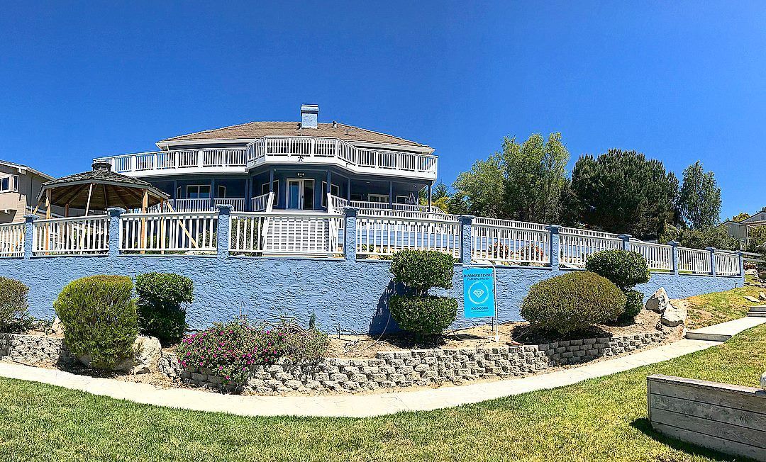 Blue mansion with multiple balconies and a white railing, overlooking a green lawn under a clear blue sky.