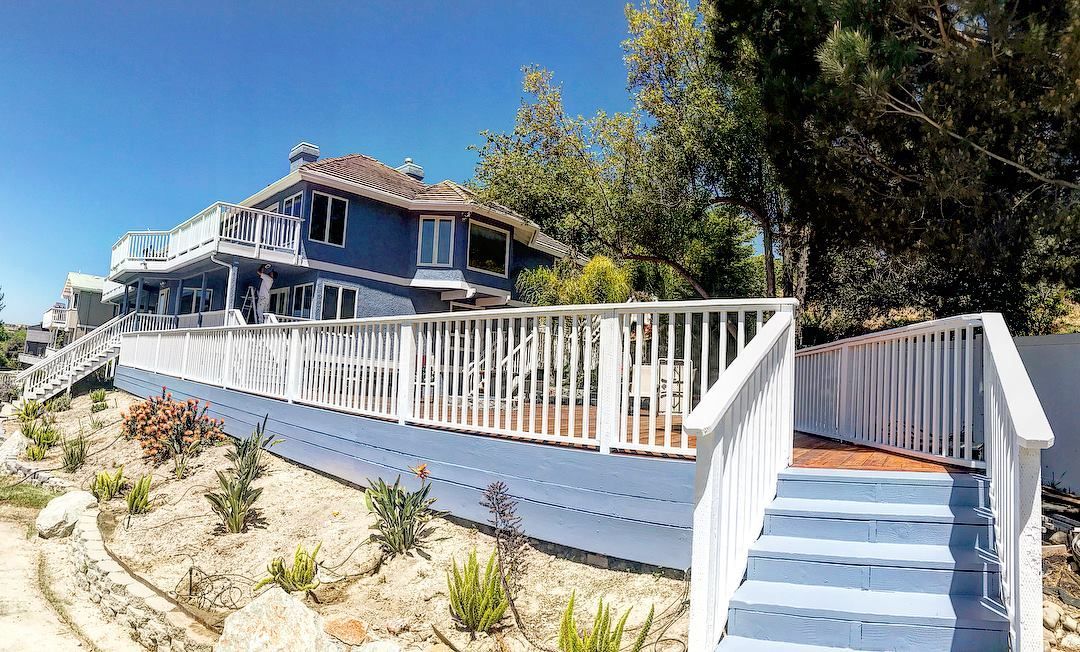 Blue house with white deck and stairs on a sunny hillside.