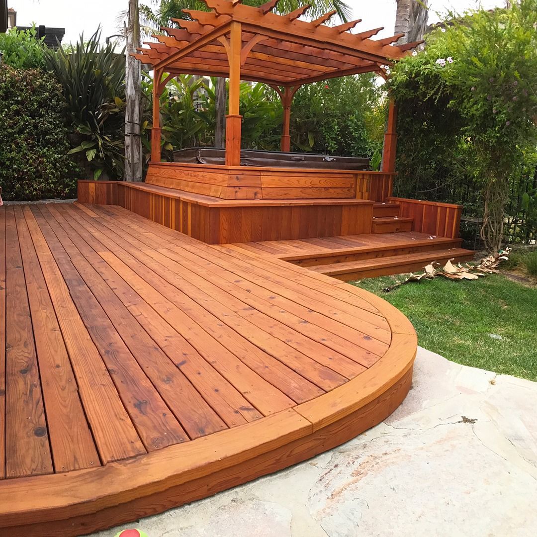 Wooden deck with built-in hot tub, pergola, and curved edge, set in a backyard with greenery.