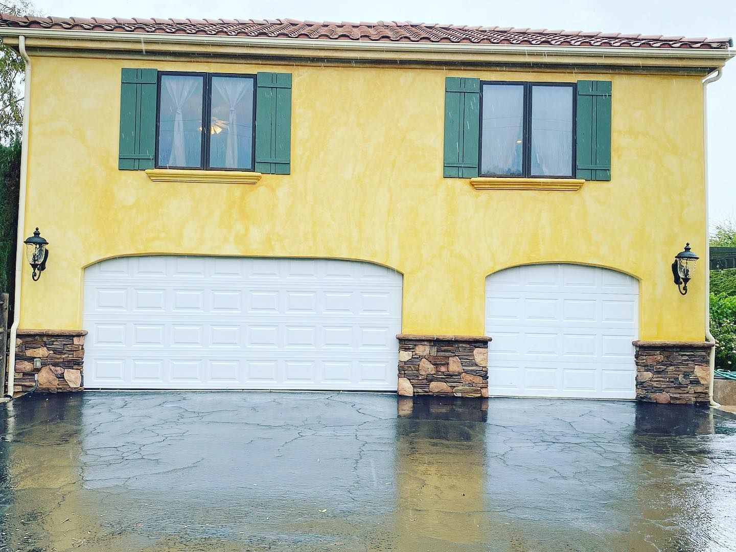 Yellow stucco building with two garage doors and rain on the driveway. Green shutters on upper windows.