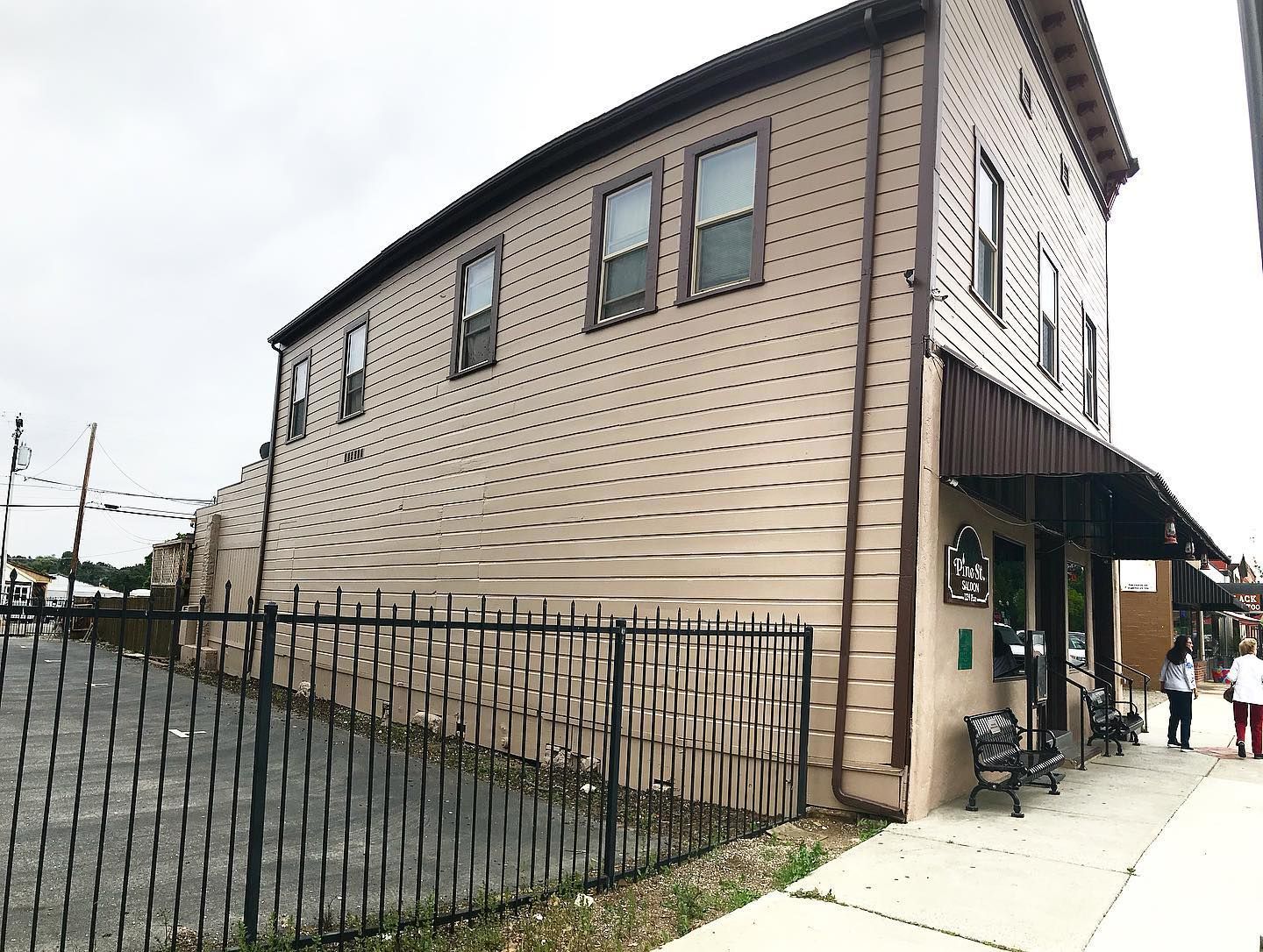 Two-story beige building with awning and black fence; people on sidewalk.