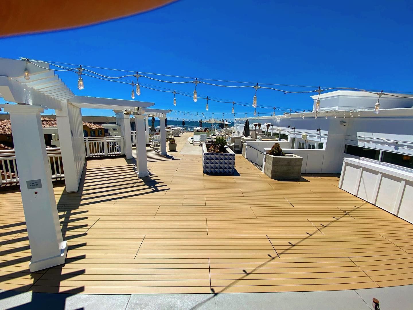 Rooftop deck with white pergolas, string lights, wooden floor, and ocean view on a sunny day.