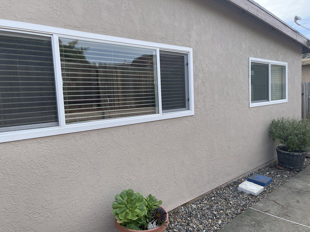 Beige stucco exterior with white-framed windows; blinds closed. Potted plants and gravel border a small yard.