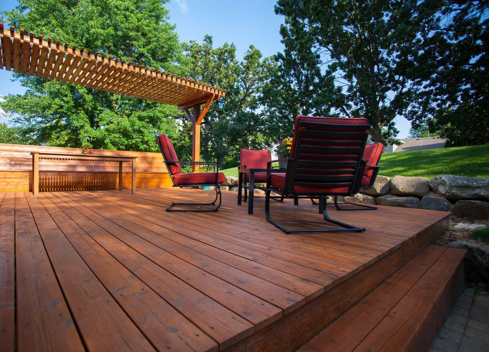 Wooden deck with red chairs and a pergola, set in a backyard with trees and a rock border.
