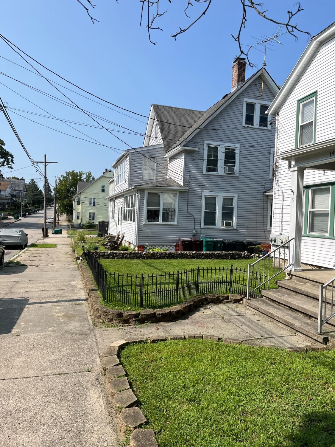 Sidewalk view of three-story houses with light-colored siding and a small grassy yard with a low brick fence in a sunny, residential neighborhood.