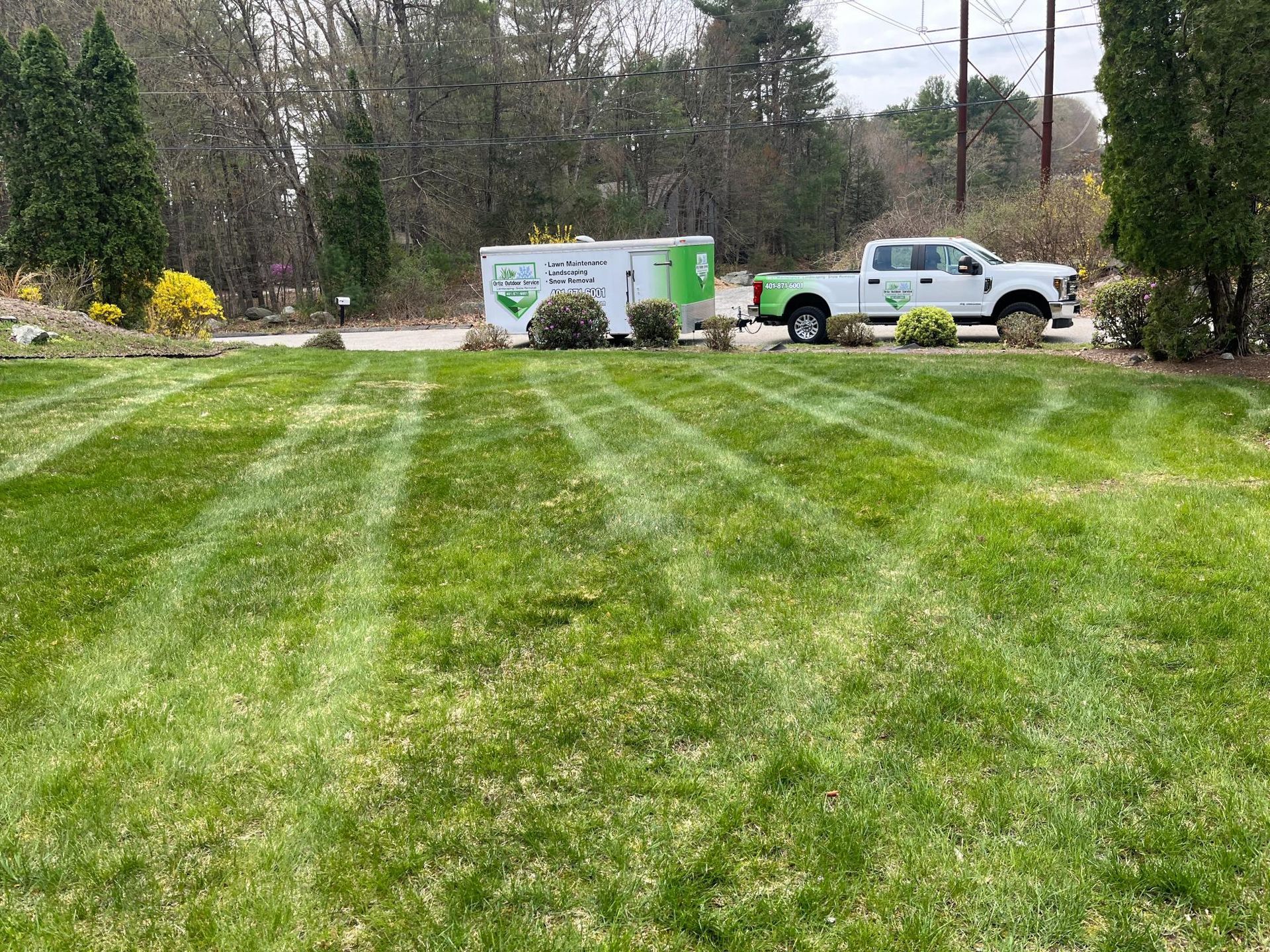 A white pickup truck with a trailer mows a lawn with a pattern, trees and building in the background.