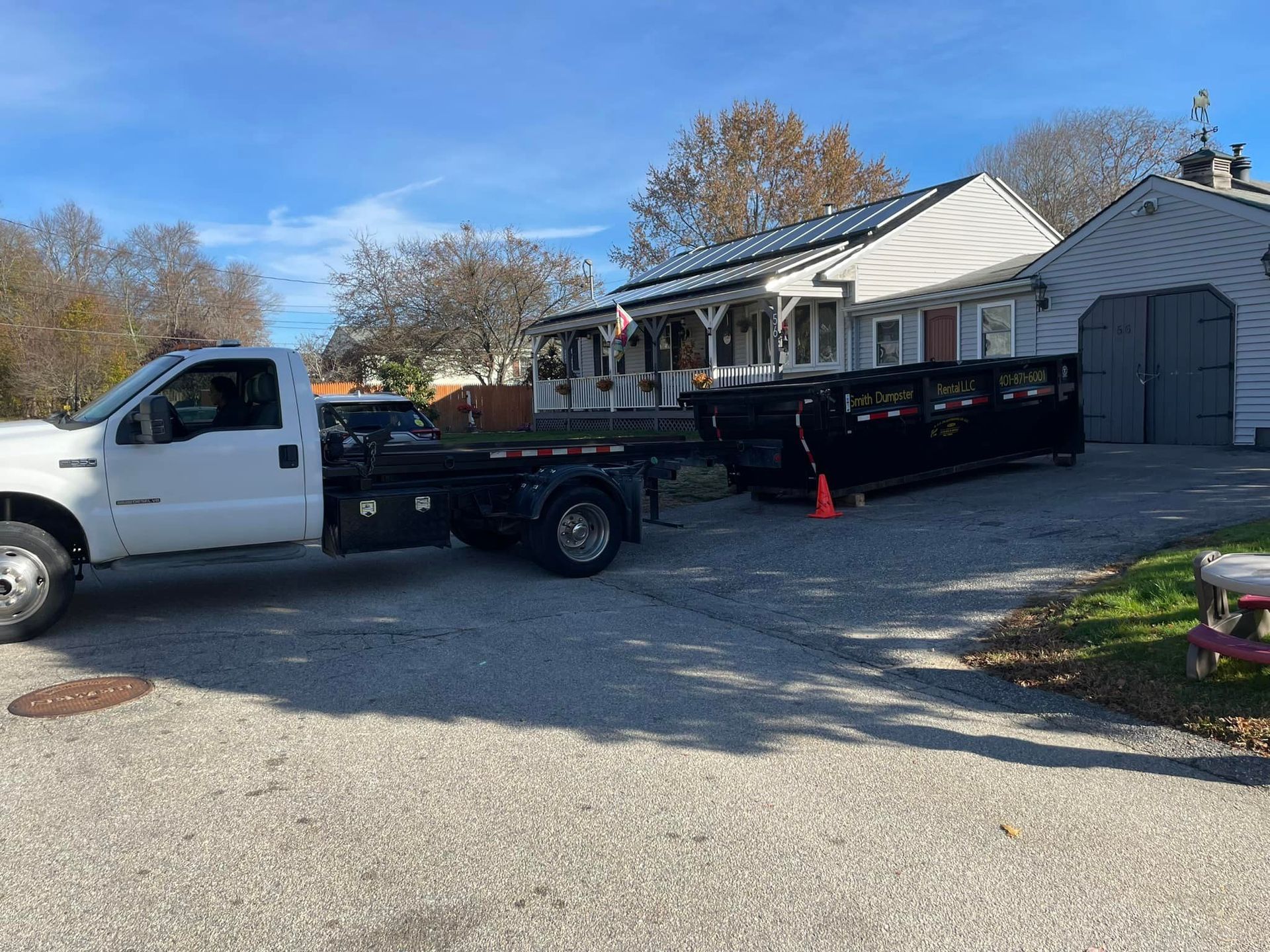 A white truck is towing a dumpster in front of a house.
