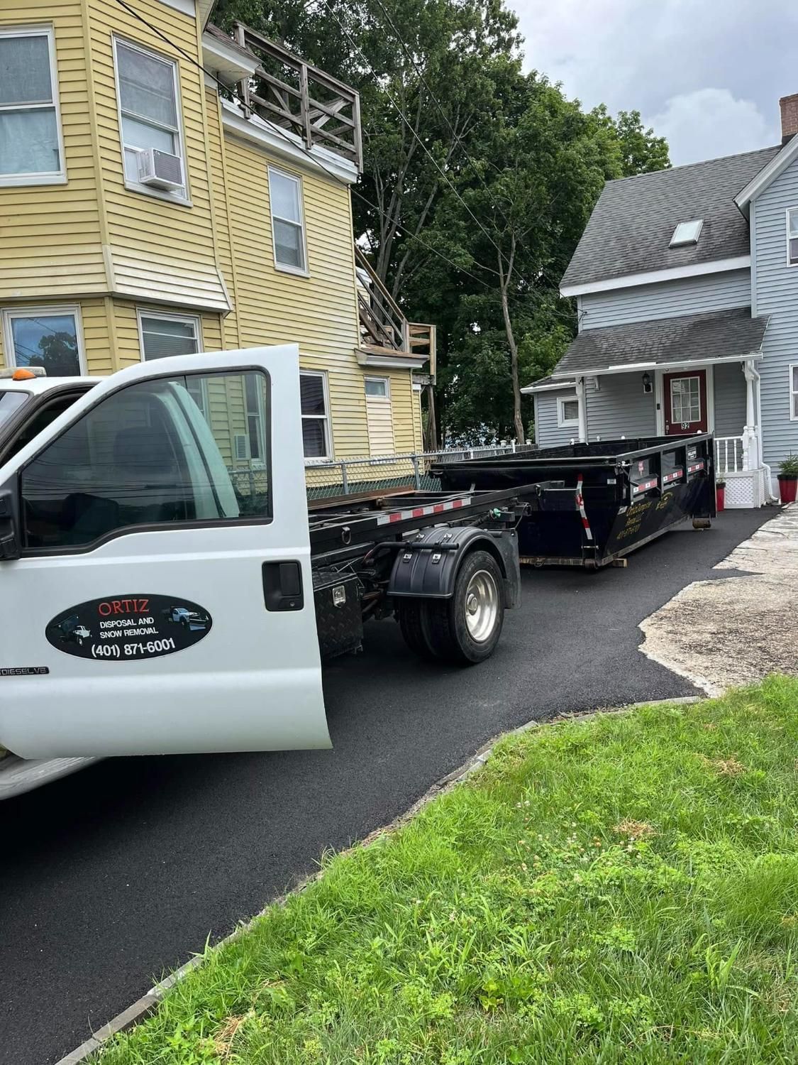 A black dumpster is being transported on a flatbed truck in a driveway. The truck is parked next to a yellow and gray house.