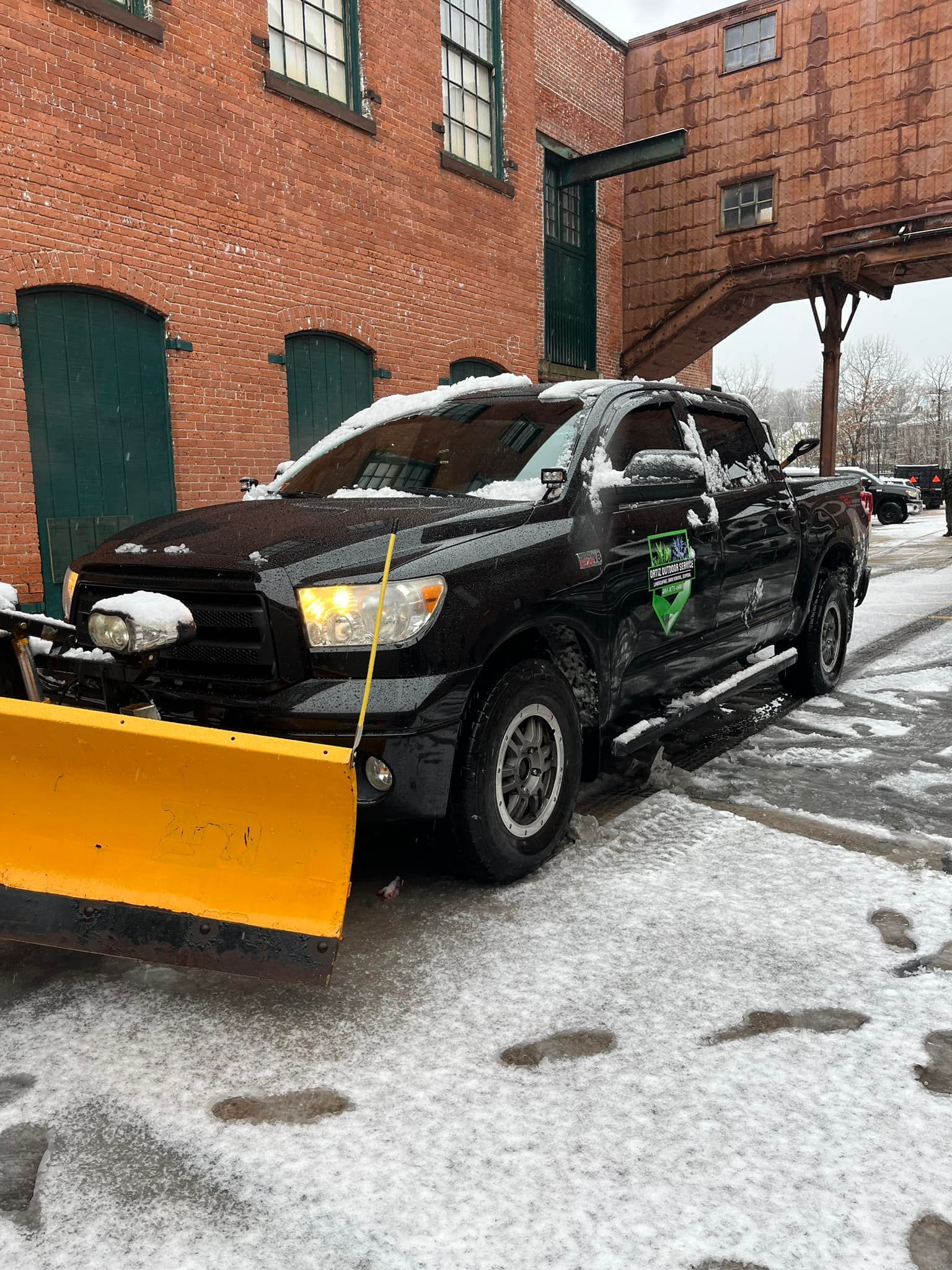 A black truck with a yellow snow plow attached to it is parked in front of a brick building.