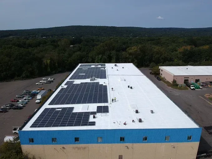 Aerial view of a large white warehouse with rooftop solar panels and a parking lot beside a wooded area.