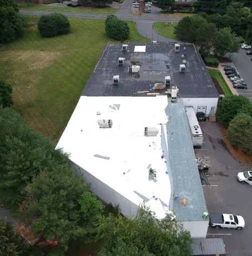 Aerial view of a flat-roof commercial building with white and black roofing, surrounded by trees and parking lots