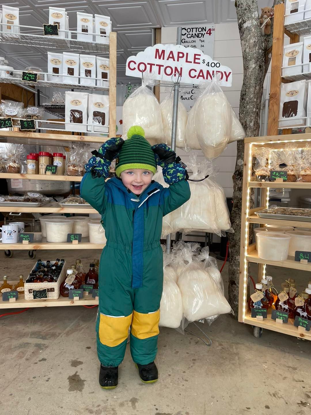 Child in green and yellow snowsuit smiles, hands on head, near cotton candy, shop shelves in background.
