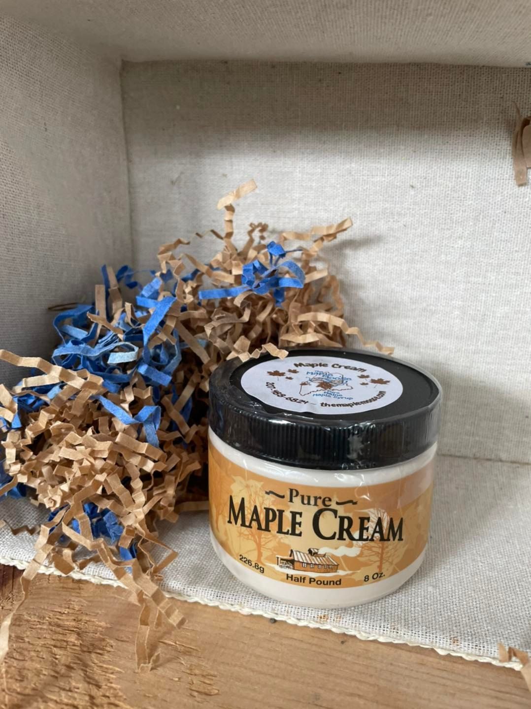 A jar of pure maple cream sits inside a wooden box with brown and blue shredded paper.