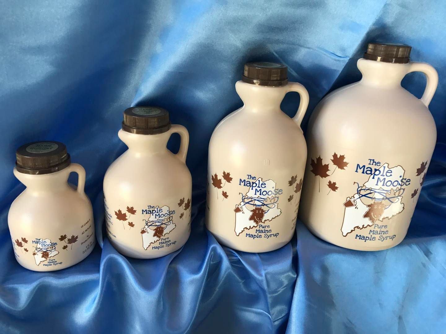 Four ceramic maple syrup jugs of varying sizes, each with a handle, cork, and Maine-themed label.