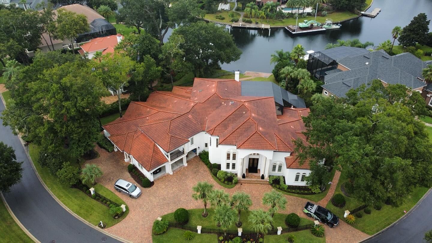Aerial view of a white mansion with a red-tiled roof, circular driveway, and waterfront access.