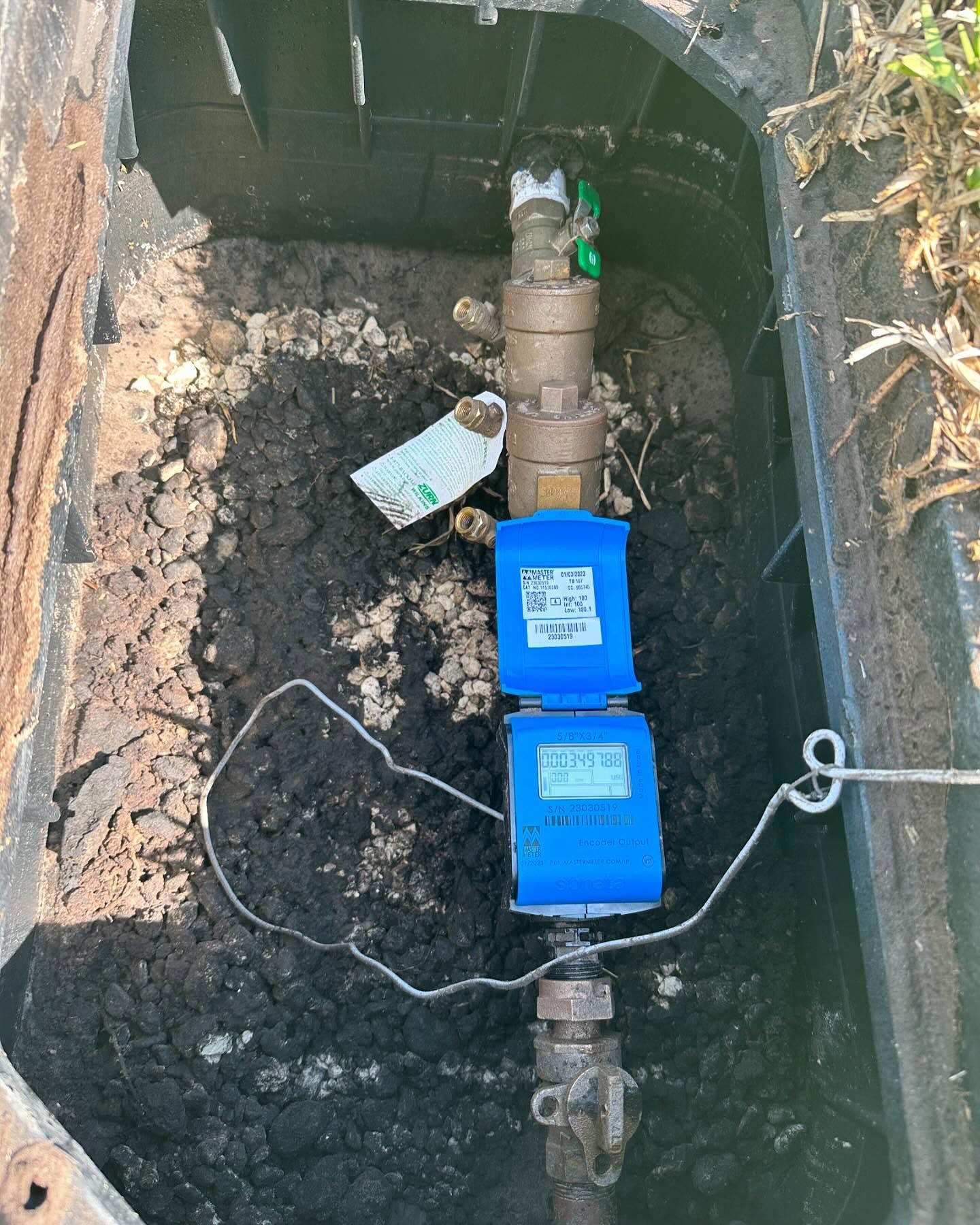 Water meter in a black plastic box. Two blue digital displays are stacked vertically on a brass pipe in dirt.