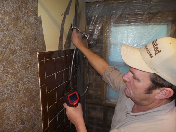 Man inspecting a tiled shower with a camera. He wears a hat and is in a bathroom setting.