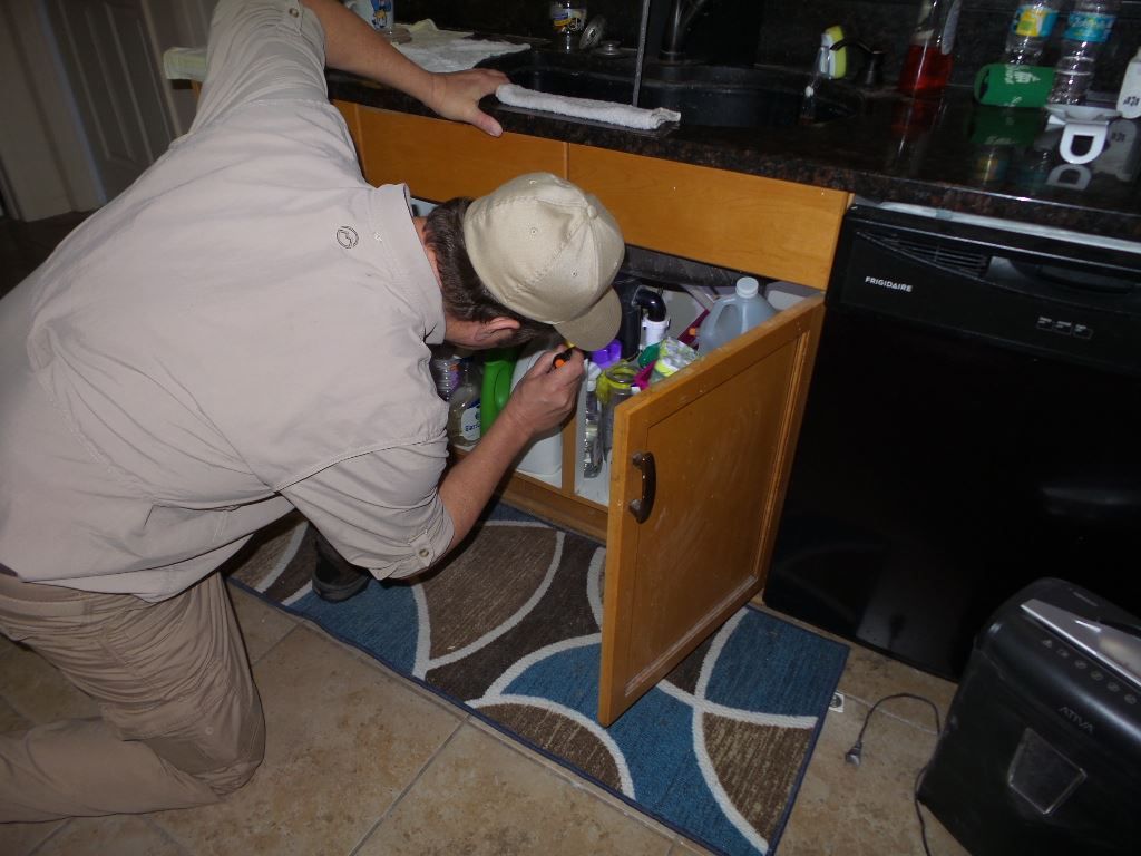 Person examining under a kitchen cabinet with open door. Beige shirt and pants, baseball cap, small rug.