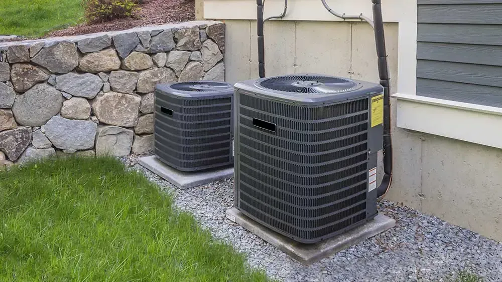 Two black air conditioning units outside, next to a building and stone wall, on concrete pads.