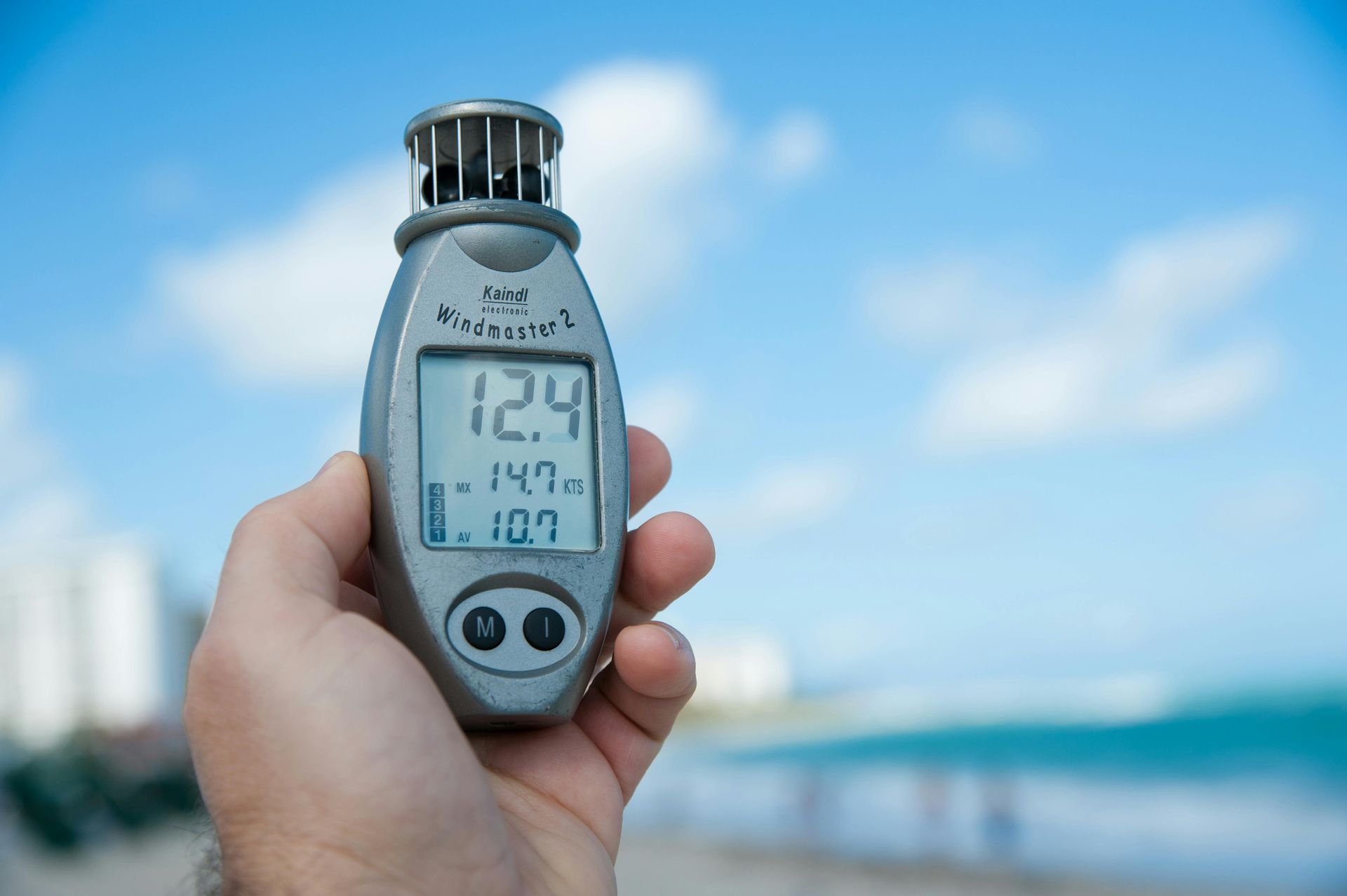 Hand holding a silver anemometer displaying readings against a blue sky, beach, and buildings.