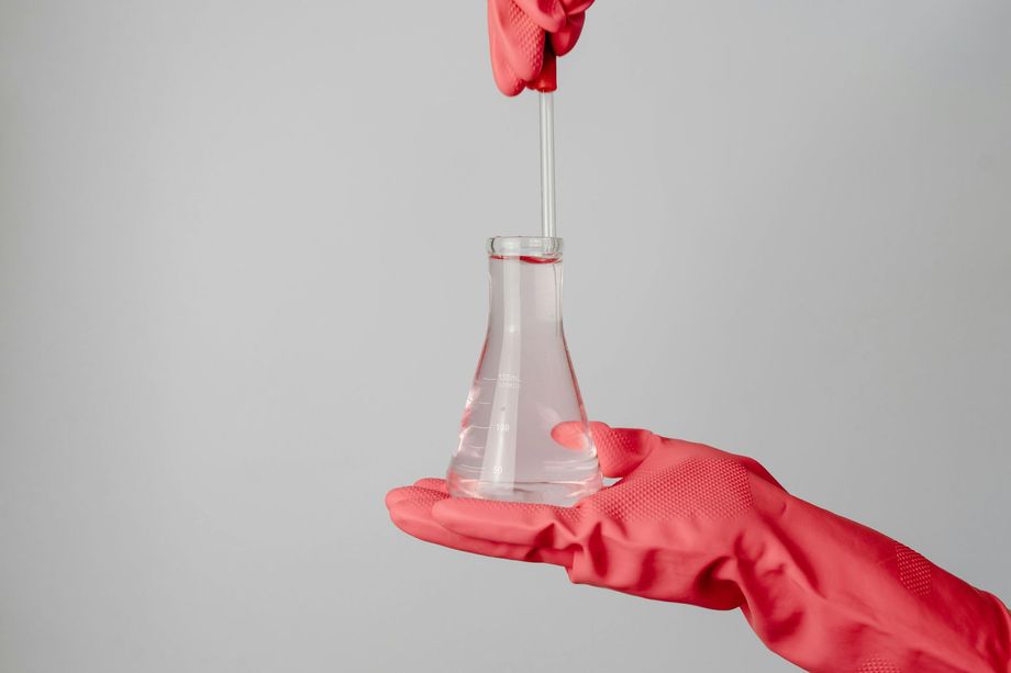 Red-gloved hands holding a flask with a liquid inside. A pipette dispenses liquid into the flask.