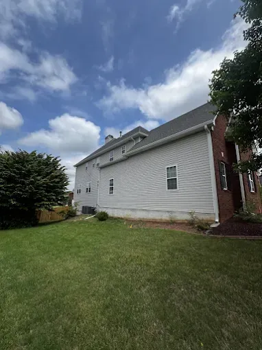 Side view of a two-story house with gray siding, brick chimney, and green grass lawn under a partly cloudy sky.