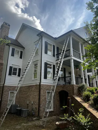 Two ladders leaning against a two-story white house with brick foundation and black shutters.