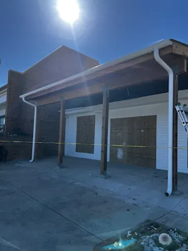 Garage with wooden posts and roof, boarded up doors, white siding, and brick.