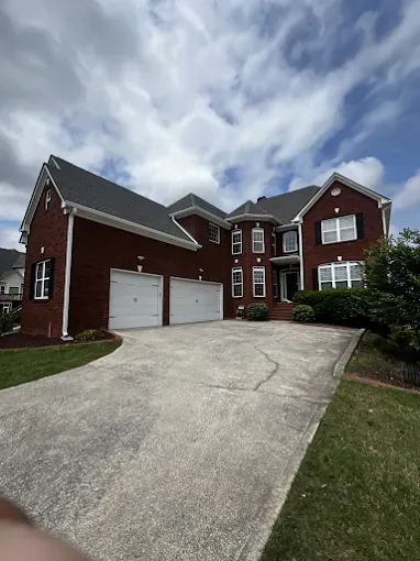 Red brick house with three-car garage, black shutters, and curved driveway under a partly cloudy sky.