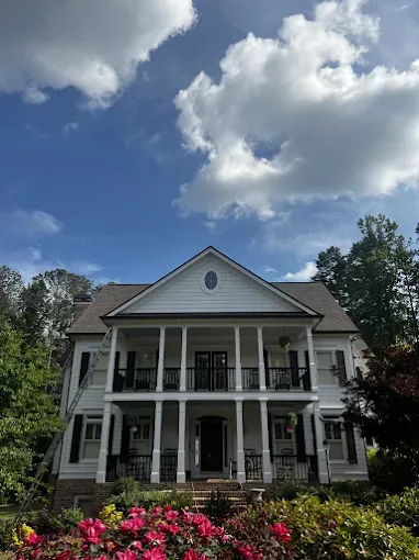 White two-story house with front porch, columns, and black shutters, set against a bright blue sky with clouds.