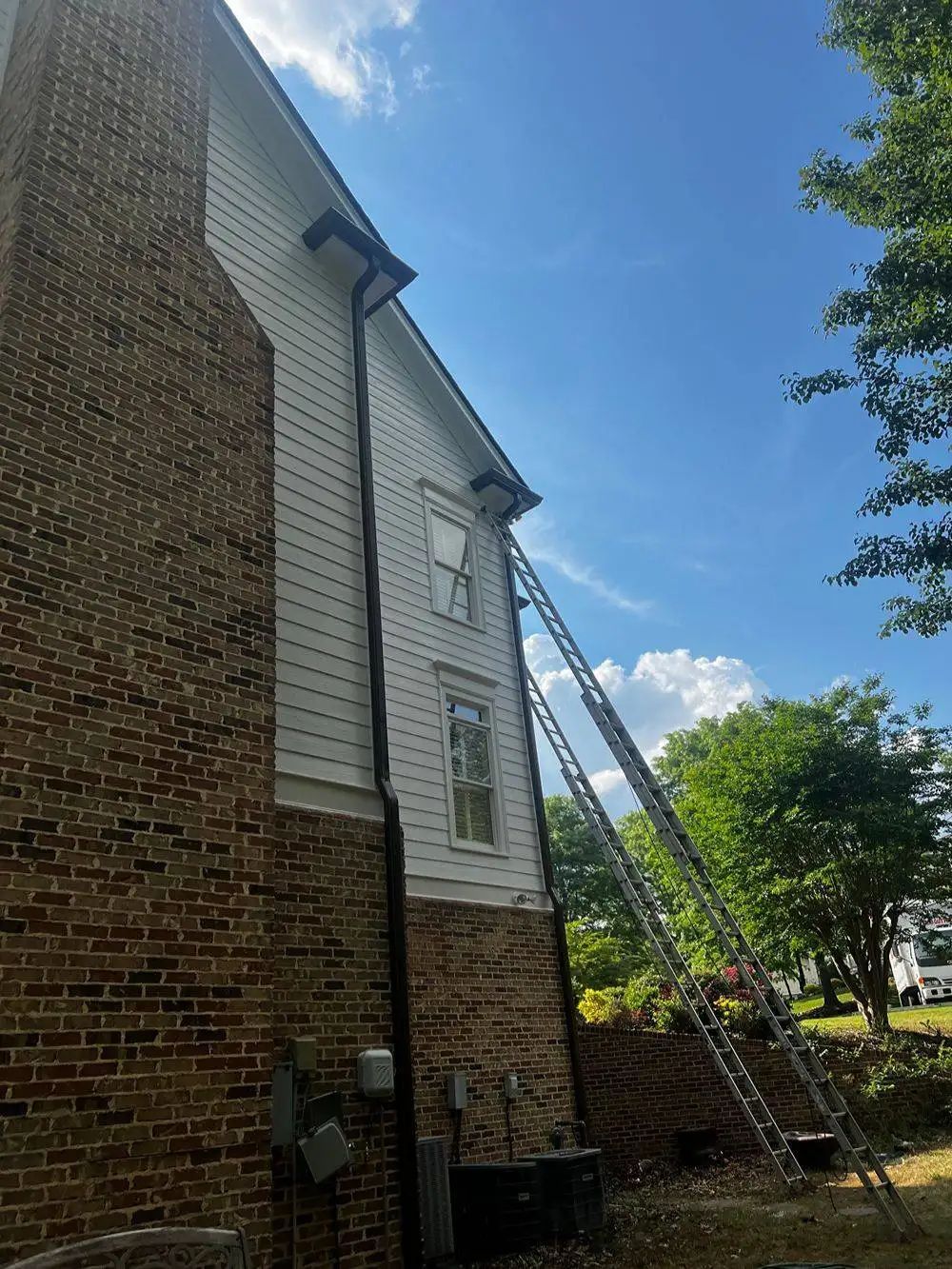 Side of a two-story house with brick base and white siding. A tall ladder leans against the house, reaching the roof.