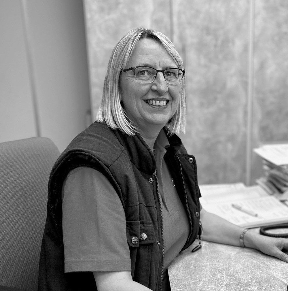 A woman wearing glasses is sitting at a desk and smiling in a black and white photo.