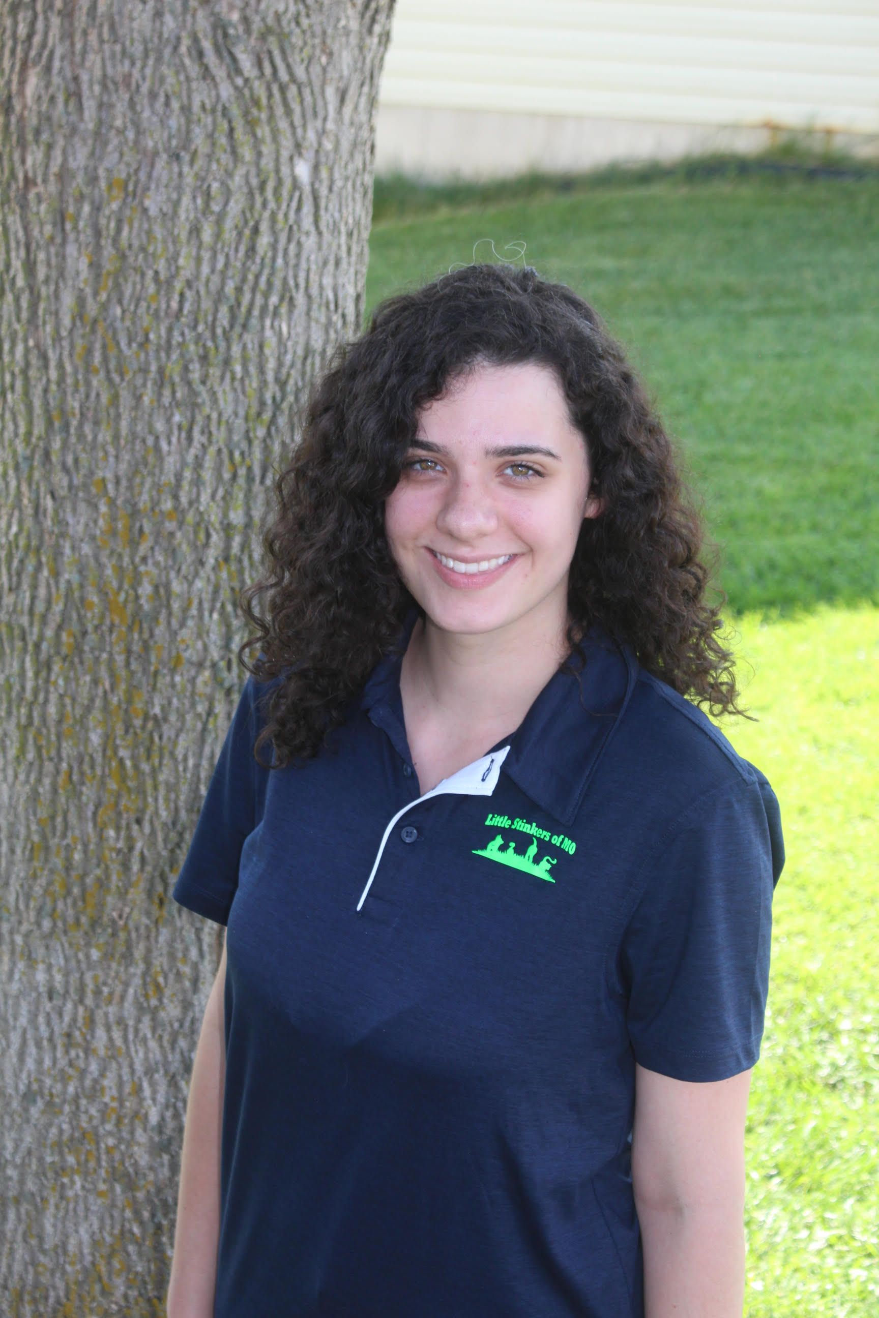 A woman with curly hair is smiling in front of a tree.
