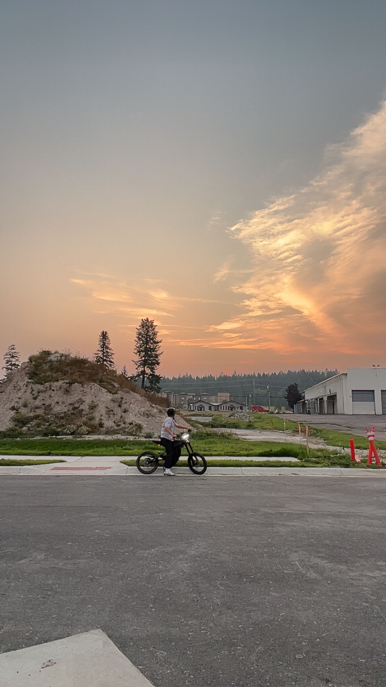 Person on a small motorcycle on asphalt road with sunset sky and construction site in background.