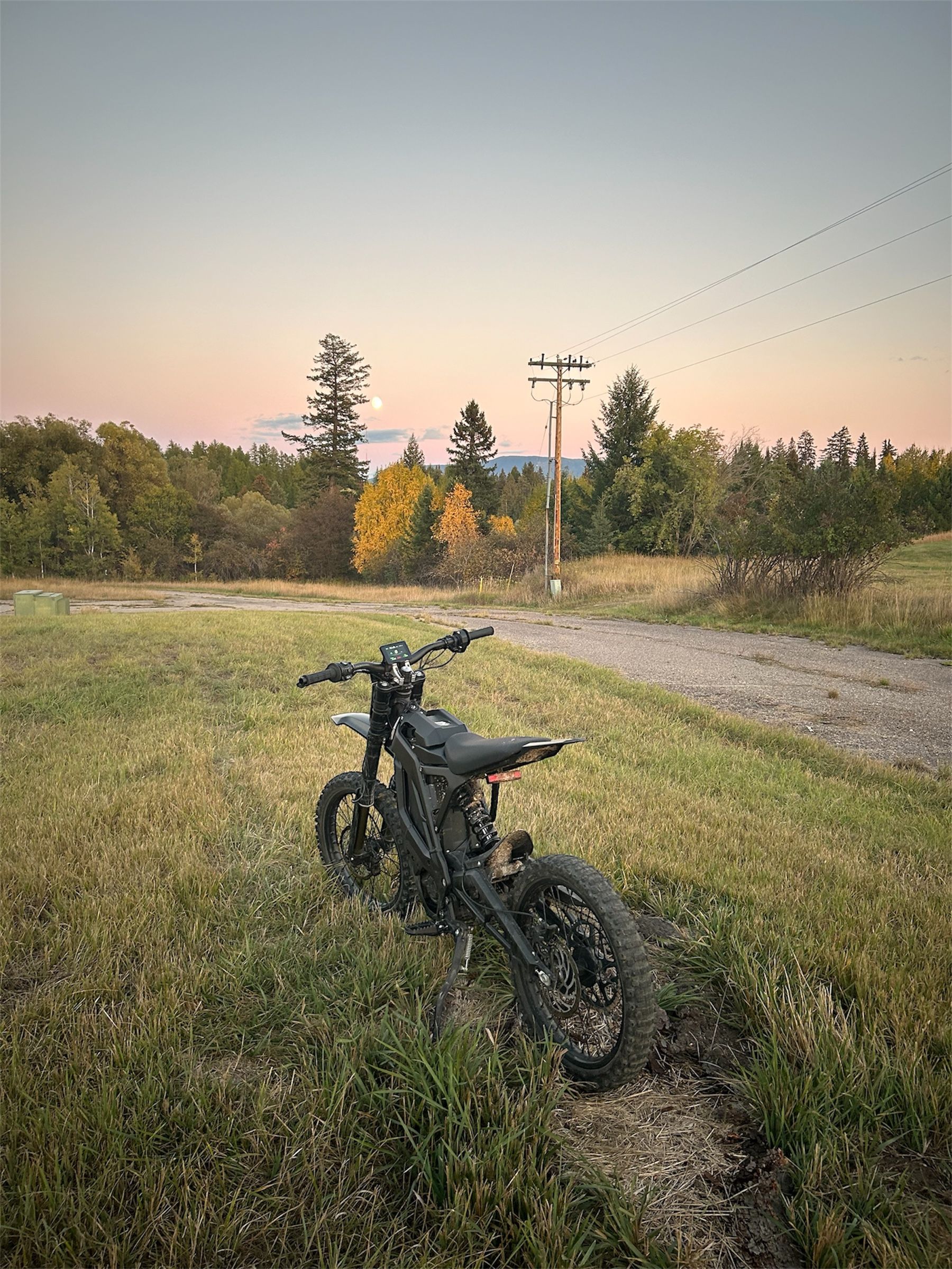 Black electric motorcycle in a field with a dirt path, under a pastel sky.