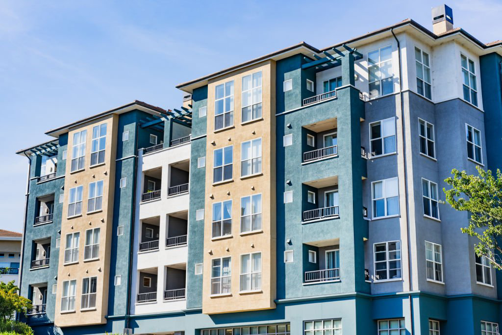 Multi-story apartment building with blue, tan, and white exterior paint under a bright blue sky.