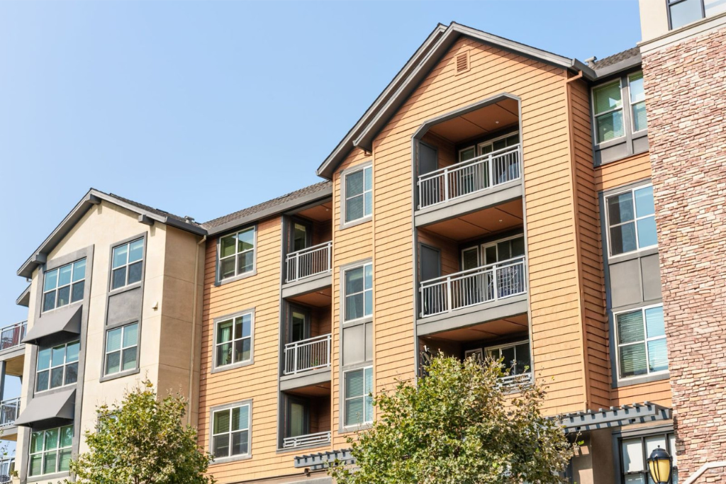 Multi-story brick apartment building with balconies, trees, and blue sky.