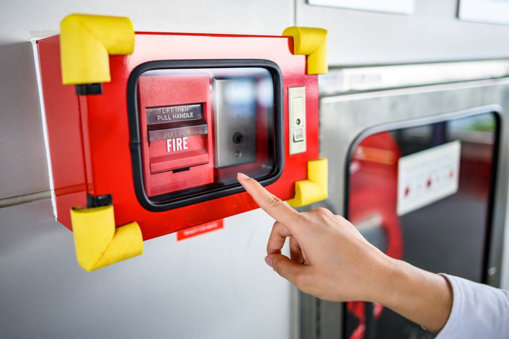 Hand presses a red fire alarm with a glass cover, mounted on a wall. Yellow corner protectors.