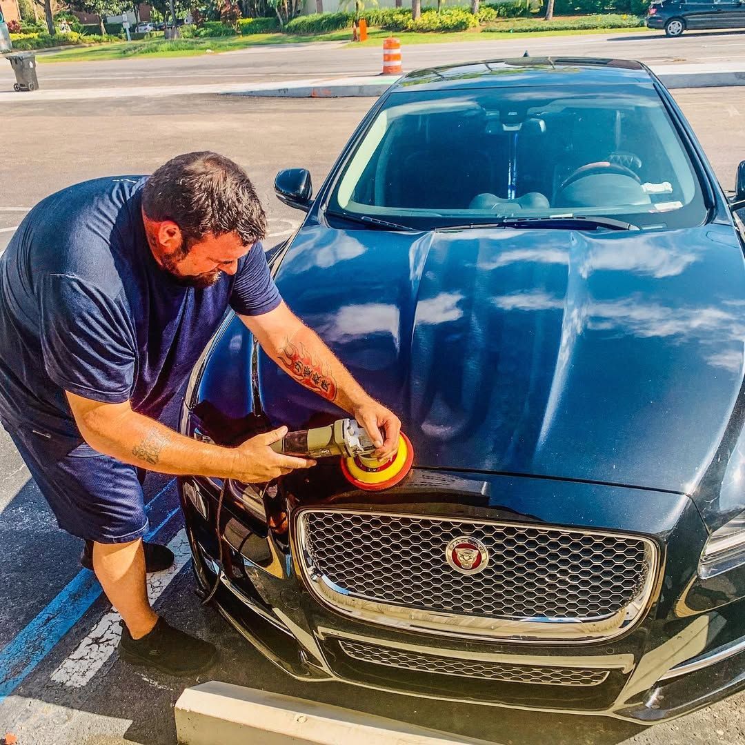 Man polishing a black Jaguar car hood with an electric buffer in a parking lot.