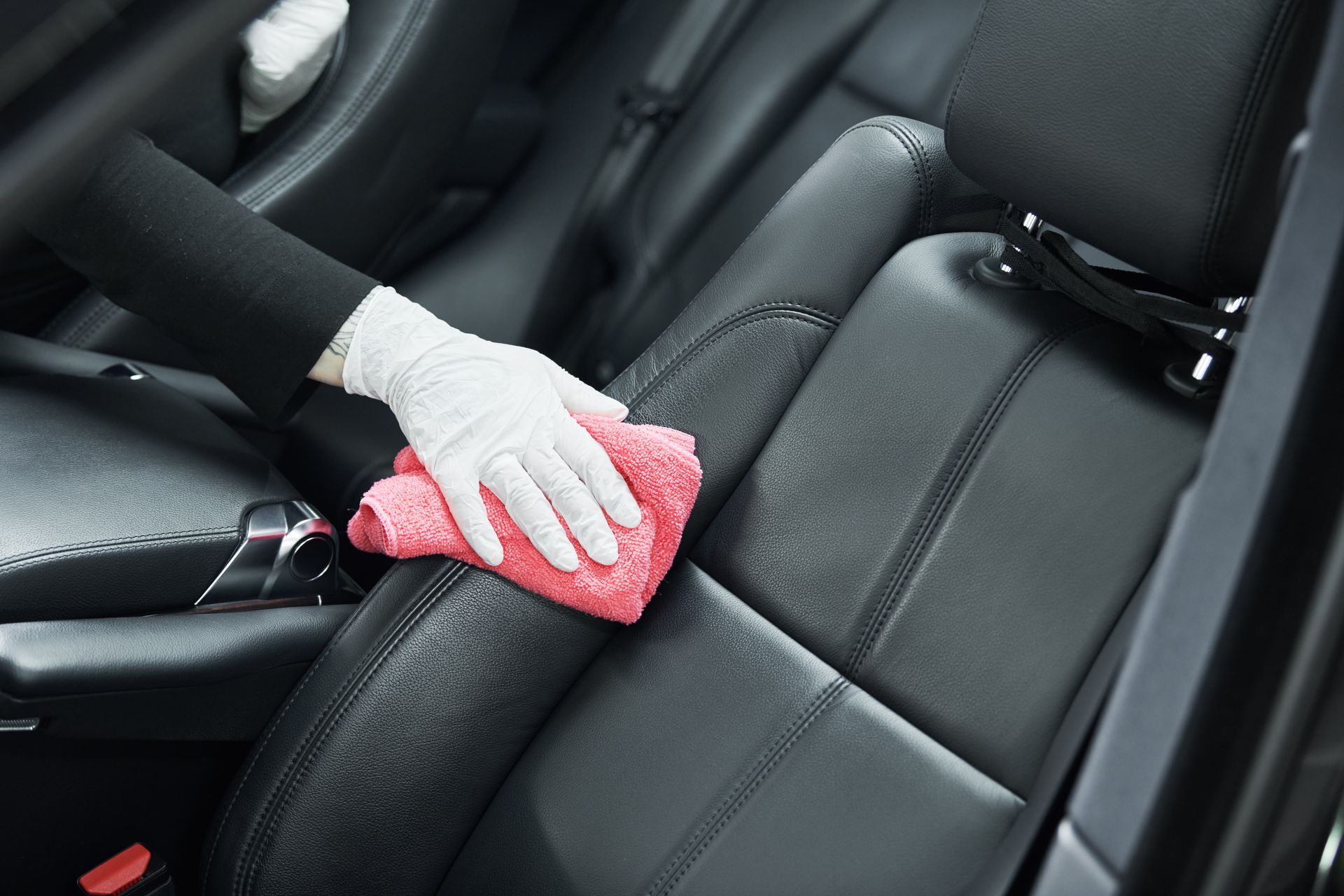 Hand in a white glove wiping a red cloth on a black leather car seat.