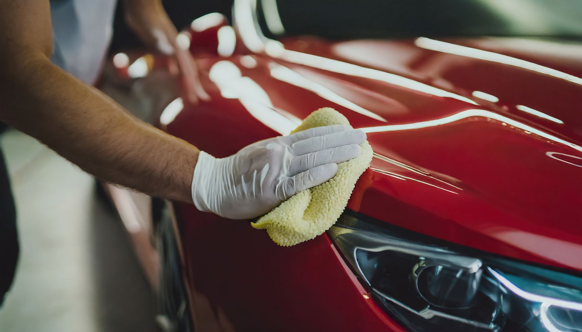 Person wearing white gloves polishing a red car with a yellow cloth.
